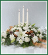 Winter white lilies, carnations and cushion pompons glow brightly from a bed of mixed evergreens. Snow-tipped pinecones, a warm sheer copper ribbon and three white taper candles adorn this elegant centerpiece. 