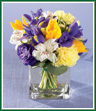 Deep purple iris and lisianthus mingle with bright yellow tulips, pastel yellow carnations and white alstroemeria. Accented by silver dollar eucalyptus and arranged in a rectangular glass vase, it's a stunning addition to any tabletop. Style of vase may vary.