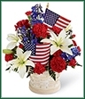 The American Glory Bouquet bursts with patriotic pride and heartfelt beauty. Blue delphinium, bright red carnations and mini carnations and brilliant white Asiatic lilies create a spectacular display arranged amongst American Flags in a round whitewash basket, creating a lovely way to celebrate this coming July 4th holiday.  
