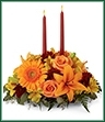 The Bright Autumn Centerpiece spins the magic of the Fall season with each sun-kissed petal to set your gathering space aglow with blooming beauty.  Two taper candles are surrounded by a gorgeous arrangement consisting of orange Asiatic Lilies, gerbera daisies, and roses with golden Peruvian Lilies, burgundy cushion poms and assorted greens to create the perfect addition to your Autumn celebration.  